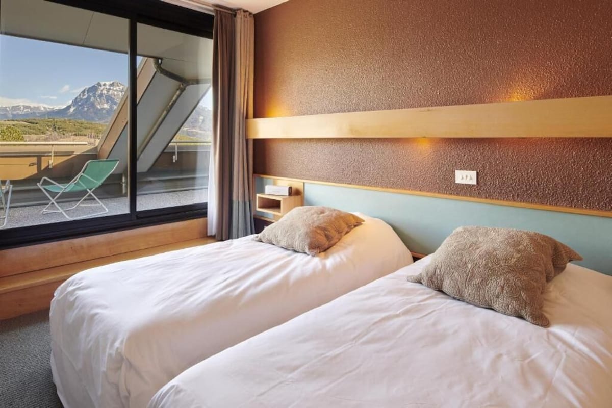 Habitación de hotel con dos camas, una ventana con vistas a montañas, una silla y una mesita.