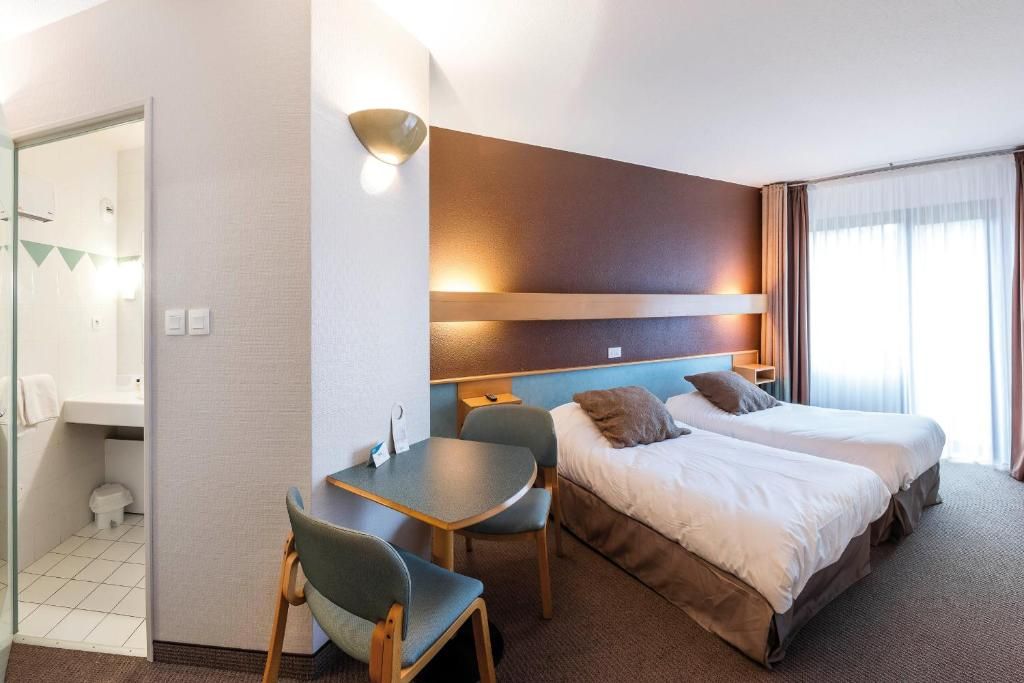 A hotel room with a bed, two chairs, a small table, a wall-mounted light, and a window with curtains.