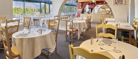 Un restaurante con mesas redondas, sillas y vistas a montañas a través de amplios ventanales.