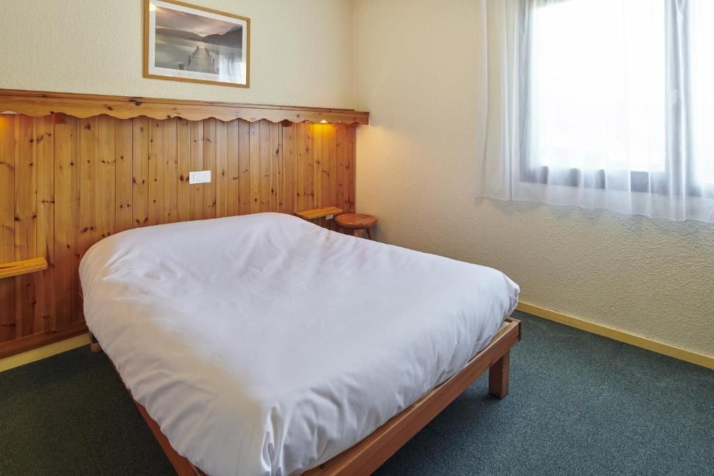 A single bed with a wooden headboard, a small stool, a framed picture on the wall, and a window with sheer curtains.