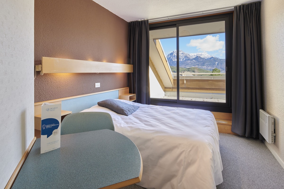 Habitación de hotel con cama, escritorio, silla y ventana con vistas a montañas, además de un cartel sobre el escritorio.