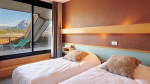 A hotel room with two beds, a window with a view of mountains, a green chair, and a wooden shelf. - Vacances Bleues Club Les Horizons Du Lac (Chorges)