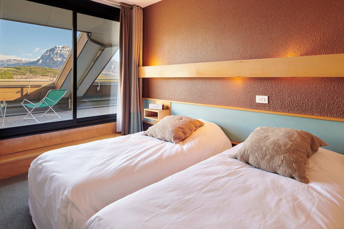 Habitación de hotel con dos camas, una ventana que ofrece vistas a las montañas, una silla verde y un estante de madera.