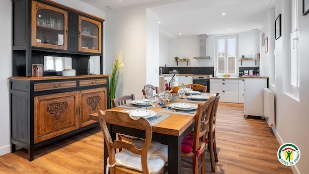 Un comedor con una mesa de madera dispuesta para cuatro personas, rodeada de sillas. Un aparador de madera oscura con puertas de cristal se encuentra a la izquierda. En el fondo, la cocina tiene armarios blancos y una encimera negra.