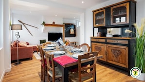 Un comedor con una mesa de madera dispuesta para una comida, rodeada de sillas. Un televisor está montado en la pared, y se ve un mueble con puertas de cristal.