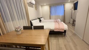 A compact hotel room with a wooden dining table, two single beds, a small kitchenette, and a large window with curtains. - Minowa Station Hideaway - Breakfast + Free Coffee & Tea (台東区)