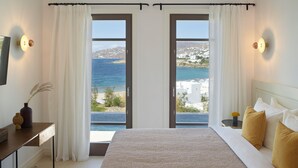 Un dormitorio con una cama grande, dos puertas que dan a un balcón con vistas al mar y edificios.