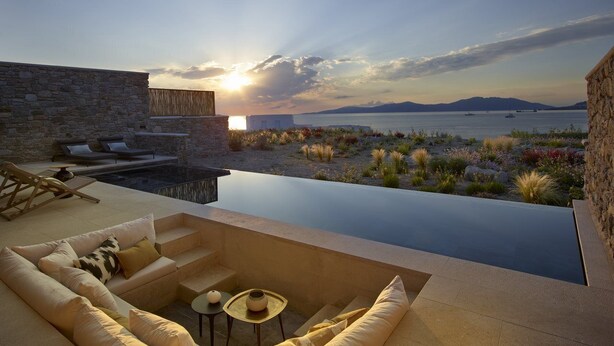 Un área de lounge exterior moderna con pared de piedra, piscina y vista al mar al atardecer.