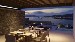 Un área de comedor al aire libre con pared de piedra, techo de madera y vista al mar y a las embarcaciones.