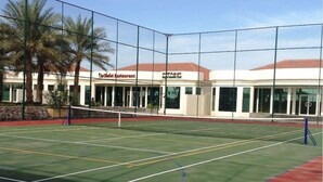 A tennis court with a net, surrounded by a fence, and a building with a sign reading "Polo Club Restaurant" in the background. - Royal Residence Resort (Umm al Quwain)
