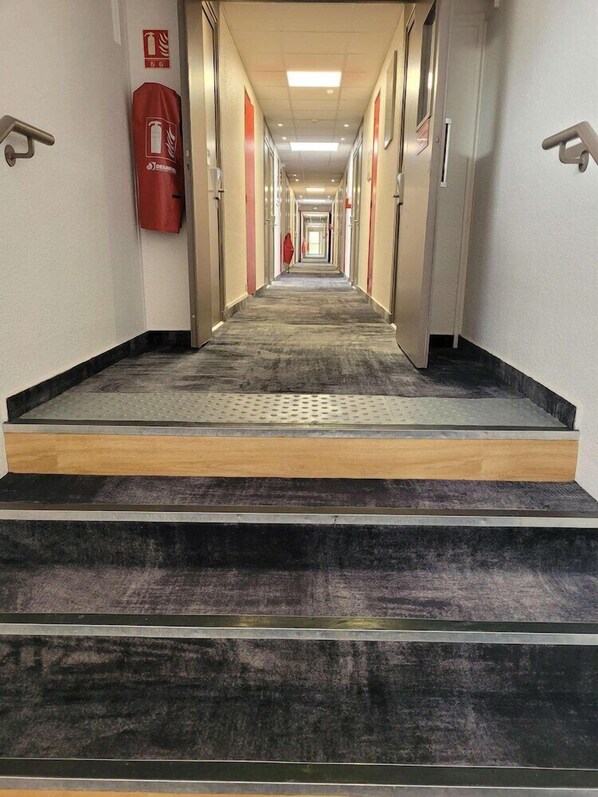 Un pasillo de hotel con piso alfombrado, un extintor en la pared y un letrero de salida de incendios.