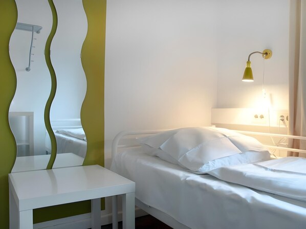 Una habitación con cama, una mesita blanca, un espejo con marco verde y una lámpara de pared.