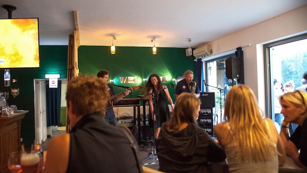 Une pièce avec un mur vert, un piano et un pied de micro. On y voit des personnes assises et debout, ainsi qu’un bar avec des verres sur le comptoir.