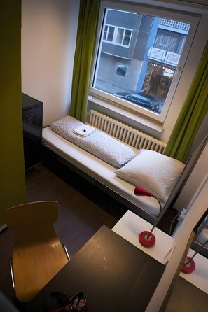 Una habitación compacta con una cama individual, un escritorio pequeño y un ventanal con vistas a la calle.