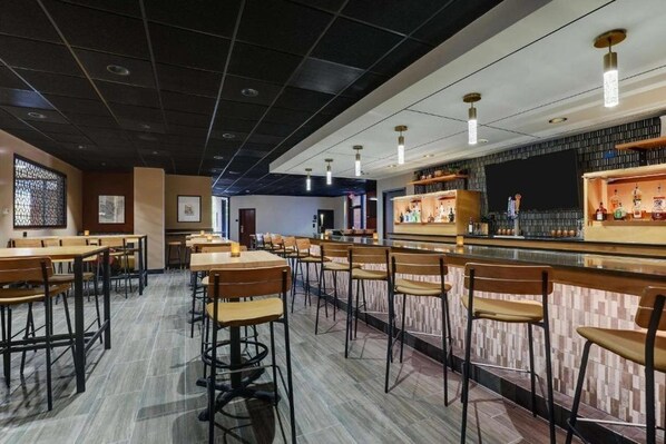 A restaurant with multiple tables and chairs, a bar area with shelves of bottles, and a large screen on the wall. - Drury Plaza Hotel Columbus Downtown (Columbus)