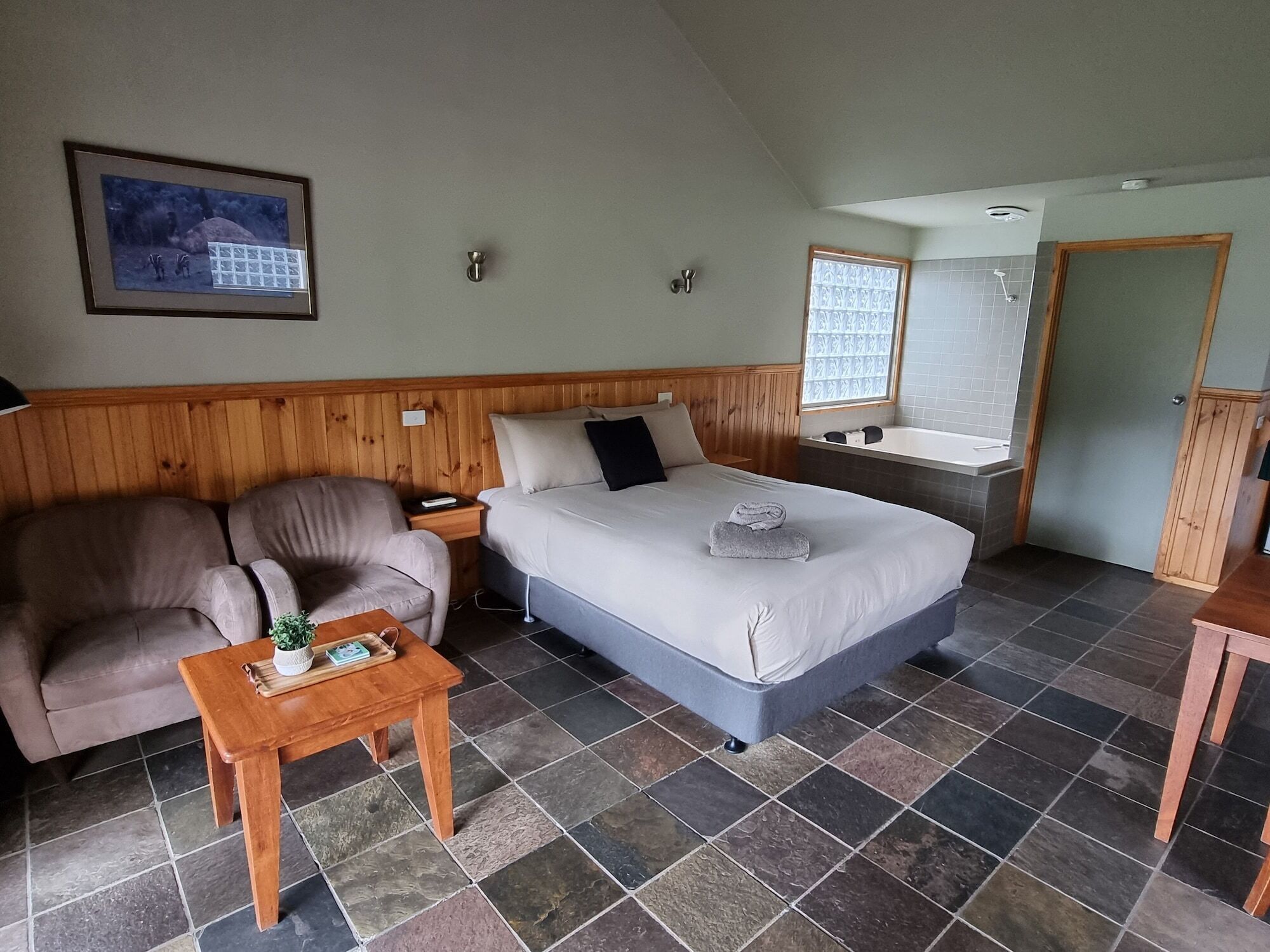 A room with a bed, a wooden headboard, a wooden bench, a small table with a plant, and a framed picture on the wall.