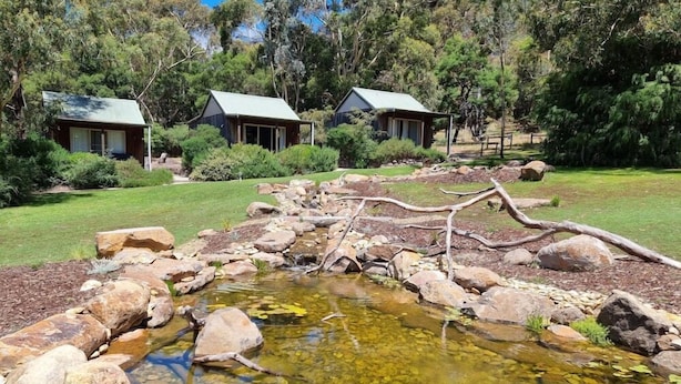 Paisagem naturalista com um pequeno lago, pedras e duas cabanas com telhados verdes.