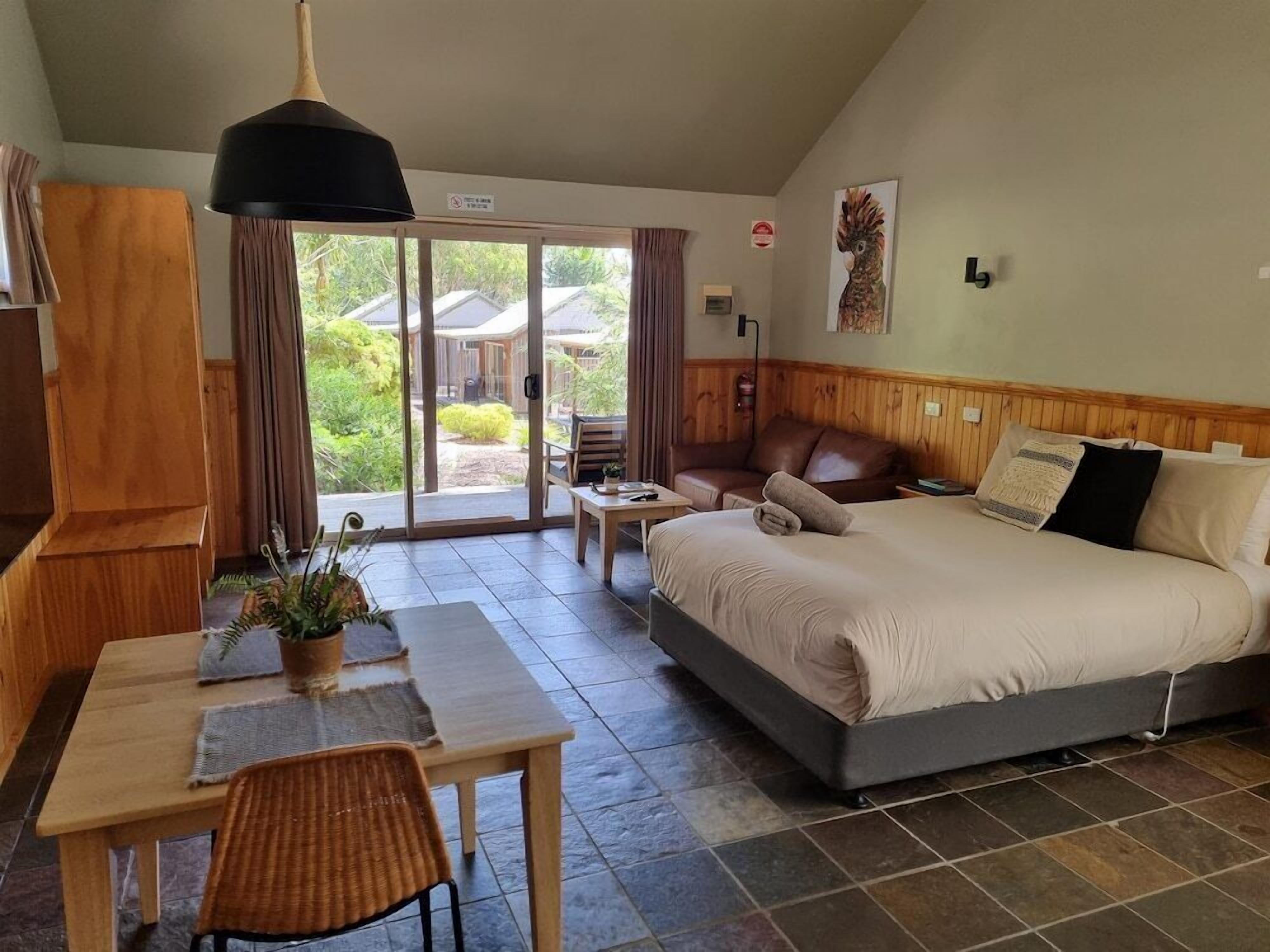 A bedroom with a bed, a desk, a chair, and a dining table. There is a sliding glass door leading to an outdoor area.
