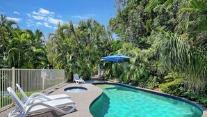 Una piscina con agua cristalina, rodeada de abundante vegetación y palmeras. Se observan sillones de descanso y un paraguas azul.