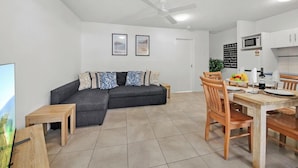 Ein modernes Wohnzimmer mit Ecksofa, Esstisch aus Holz und Stühlen. Es gibt einen Deckenventilator, eine Küchenzeile mit Schränken und Geräten sowie gerahmte Bilder an der Wand.