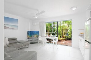 Un salon moderne avec un canapé d’angle gris, un coin repas avec des chaises blanches, un ventilateur de plafond et une grande fenêtre donnant sur un paysage verdoyant.