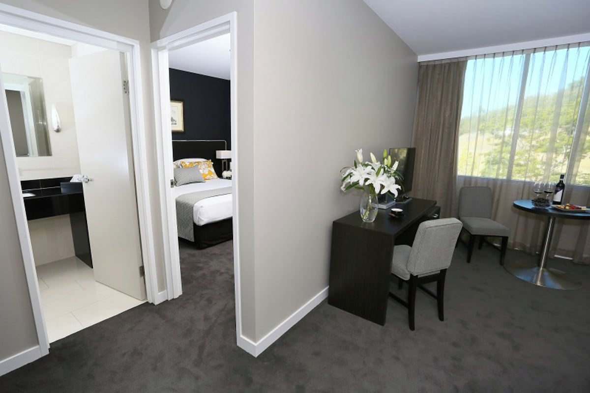 A hotel room with a bed, a desk, a chair, a small table, and a view of the outdoors.