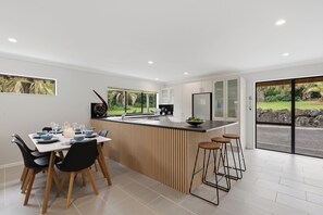 A modern kitchen with a central island, bar stools, and a dining area with chairs. - Clay Cottage your close to town country escape. (Waimauku)
