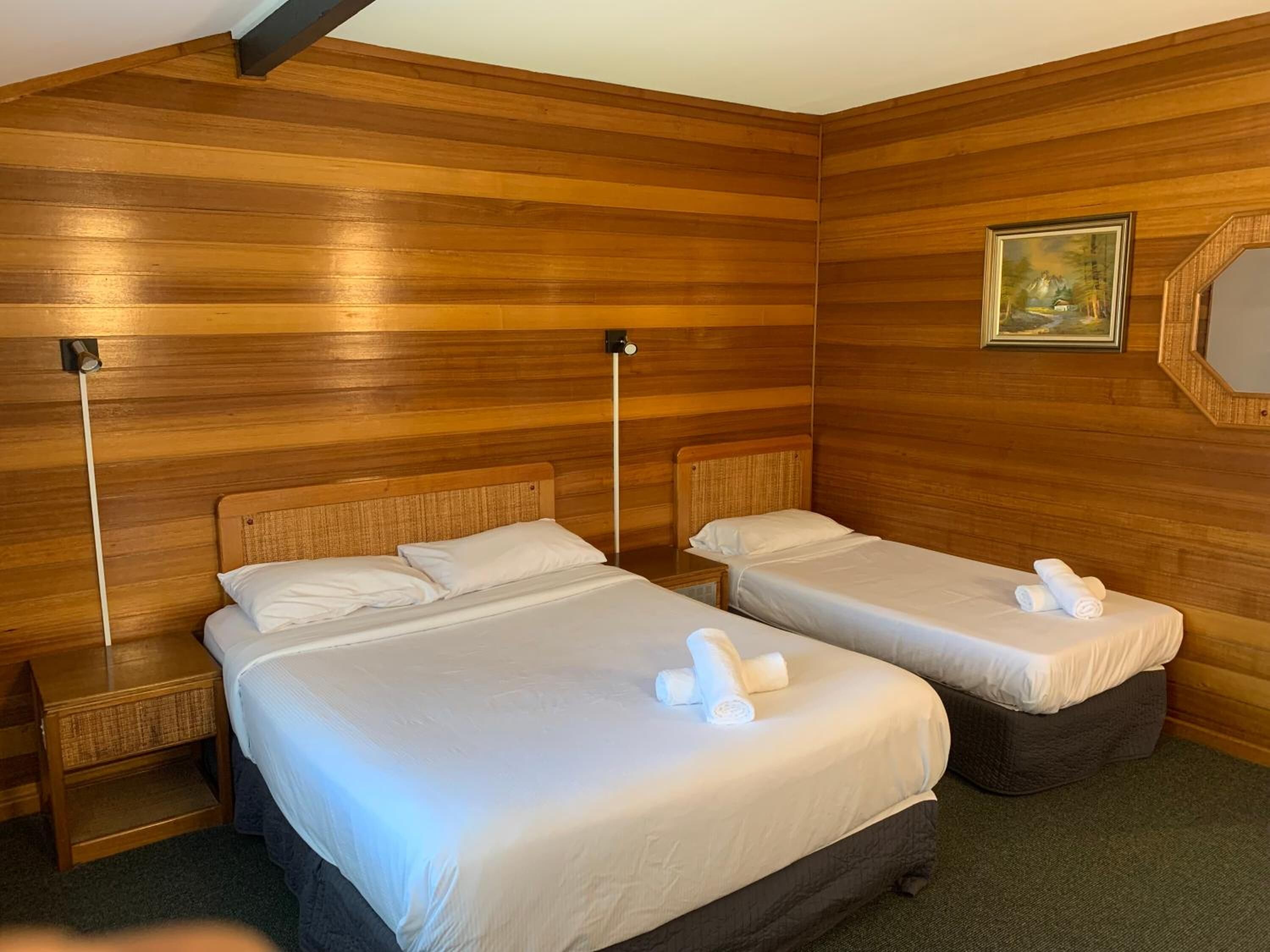 A room with two beds, each with a wooden headboard, a nightstand, and a wall-mounted lamp.