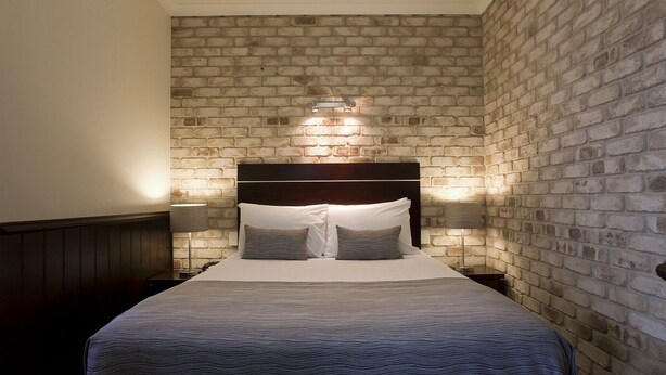 Quarto com parede em padrão de tijolos, cabeceira escura e duas luminárias de apoio.