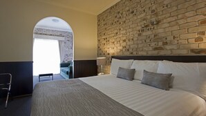Quarto de hotel com parede em tijolos, uma cama grande com roupa de cama estampada, uma mesa e uma cadeira.