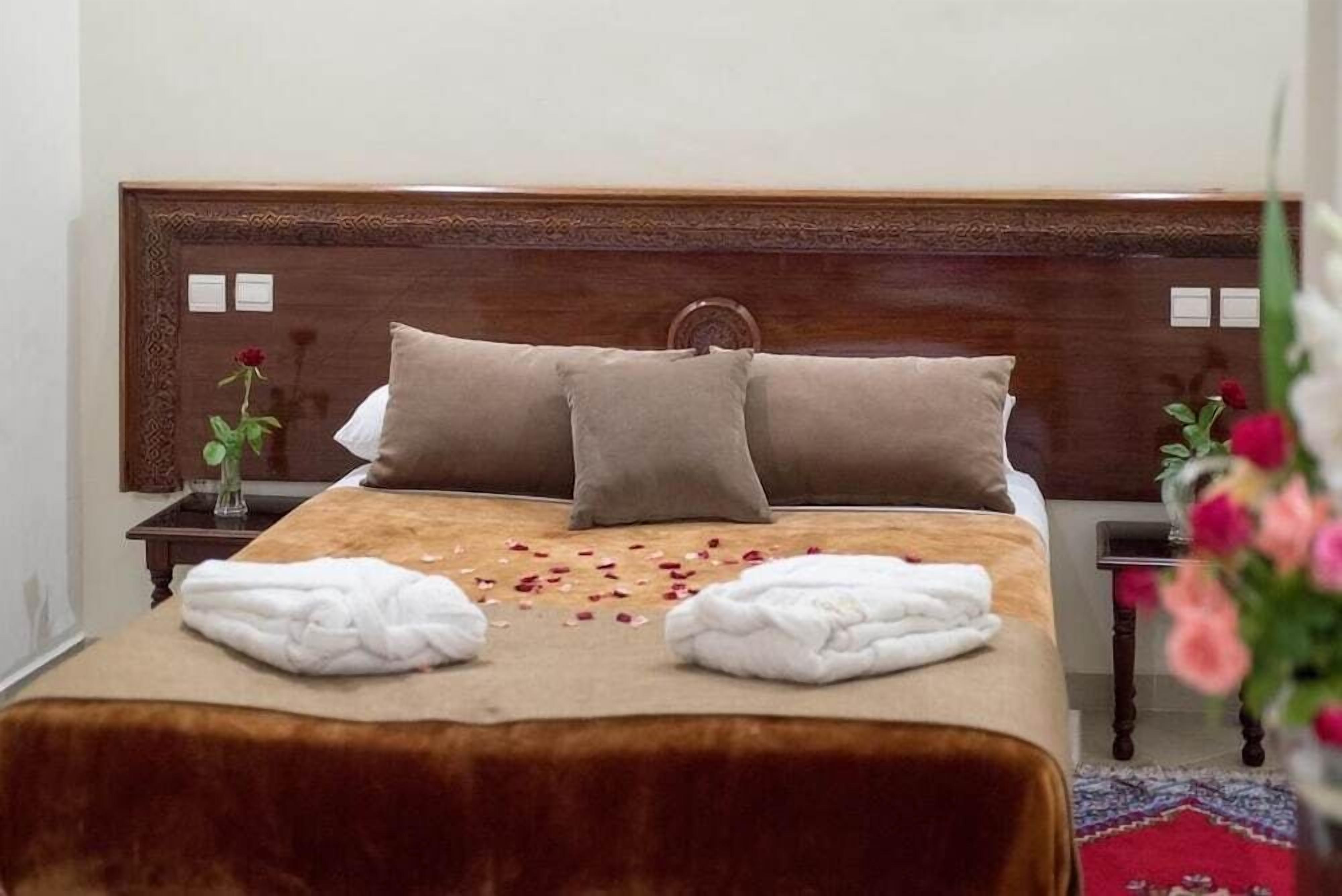 Una cama bien hecha con un cabecero marrón, dos almohadas y una colcha color beige. Hay dos toallas blancas dobladas sobre la cama y un jarrón con una sola rosa roja sobre una mesita de noche junto a la cama.