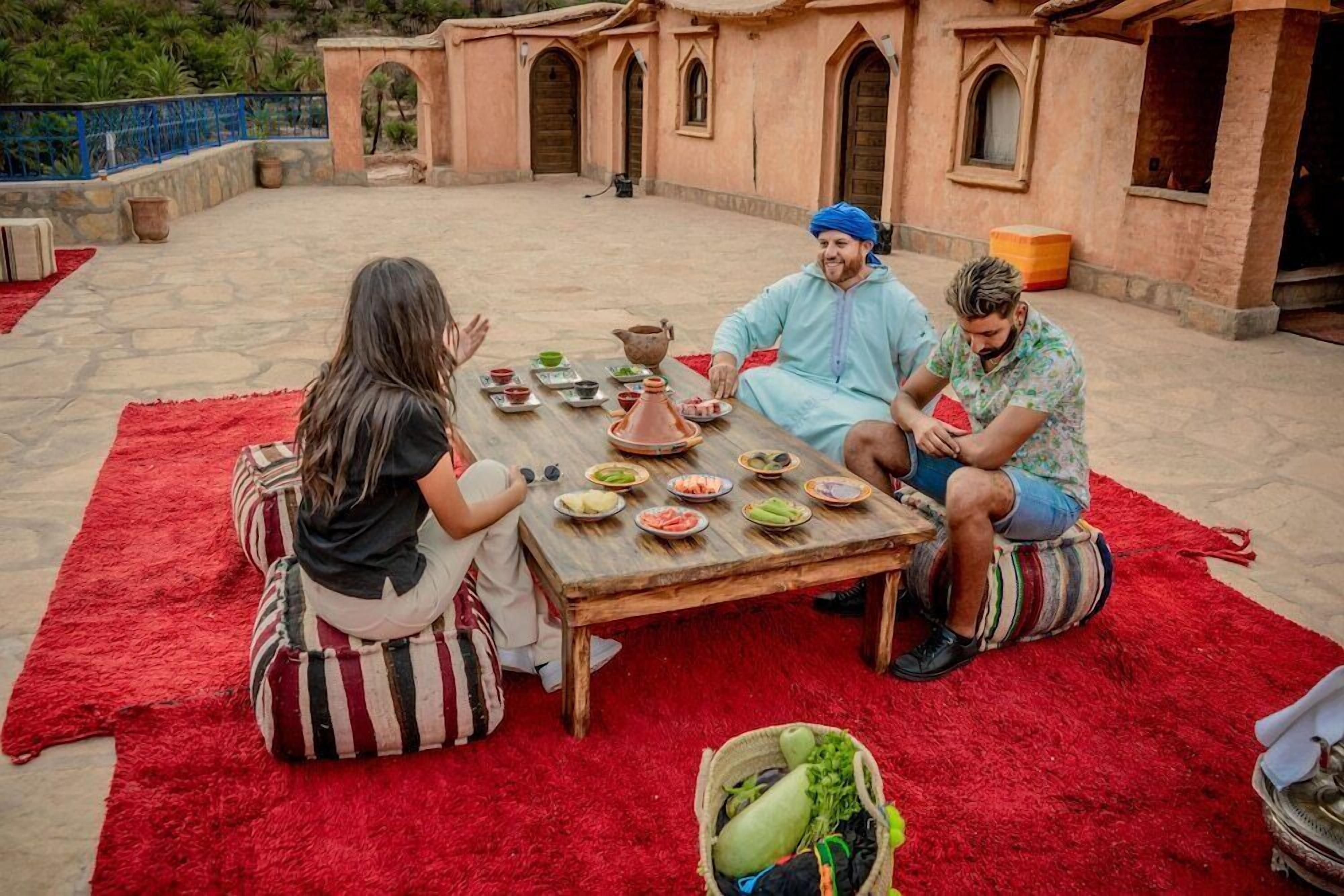 A group of people sitting on a red carpeted floor around a wooden table with various dishes and a pot, in an outdoor setting with a building in the background.