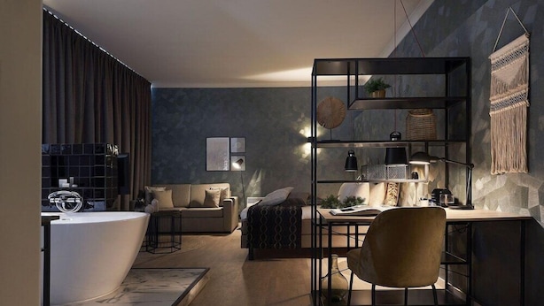 A modern living room with a built-in bathtub, a sofa, a coffee table, and a desk with a chair. There is a bookshelf with decorative items and a macrame wall hanging. - Best Western Plus Hotel Atlant (Genk)