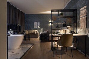 A modern living room with a built-in bathtub, a sofa, a coffee table, and a desk with a chair. There is a bookshelf with decorative items and a macrame wall hanging. - Best Western Plus Hotel Atlant (Genk)