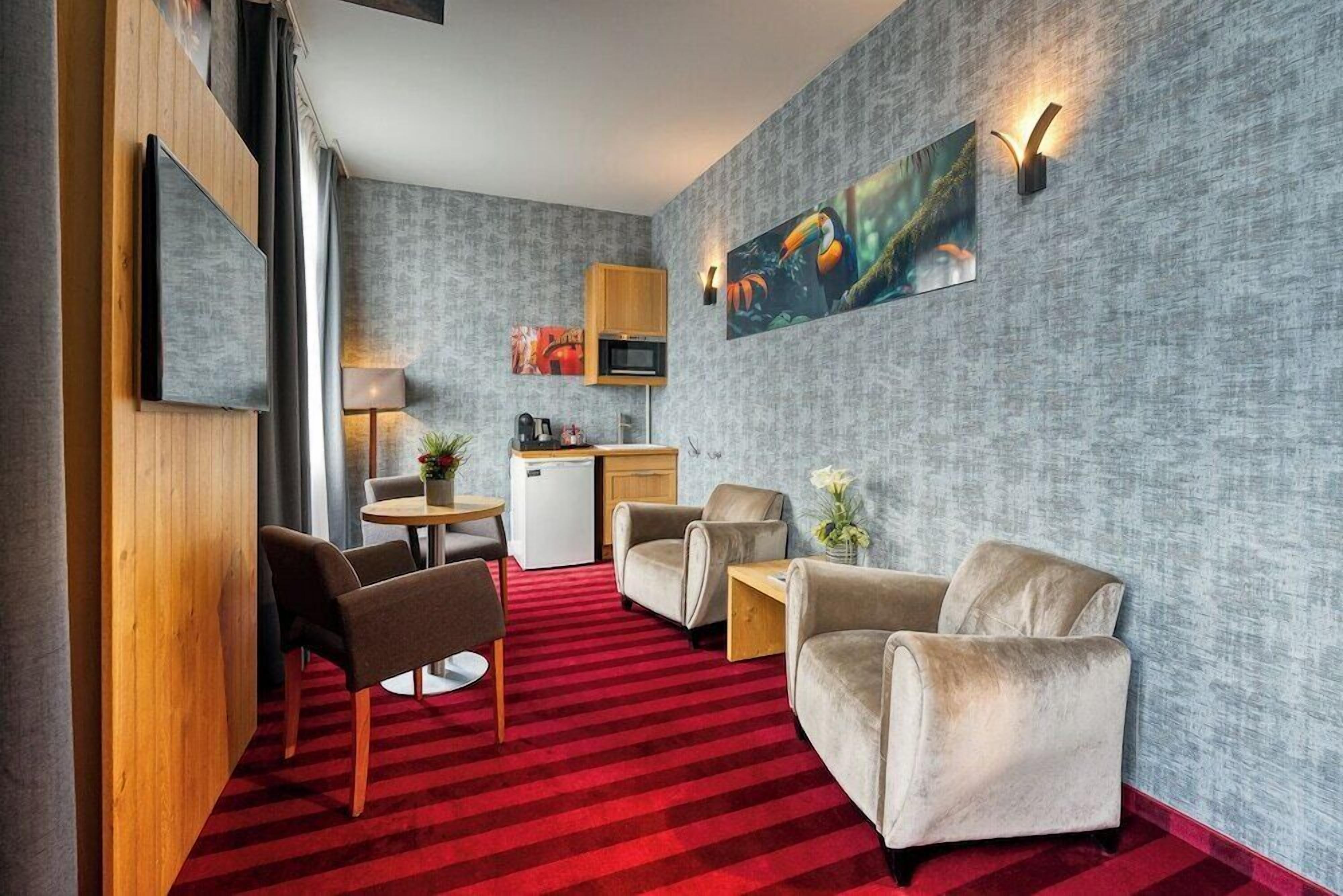 Habitación de hotel con alfombra roja, un panel de madera, kitchenette y un área de descanso con dos sillones y una mesita.