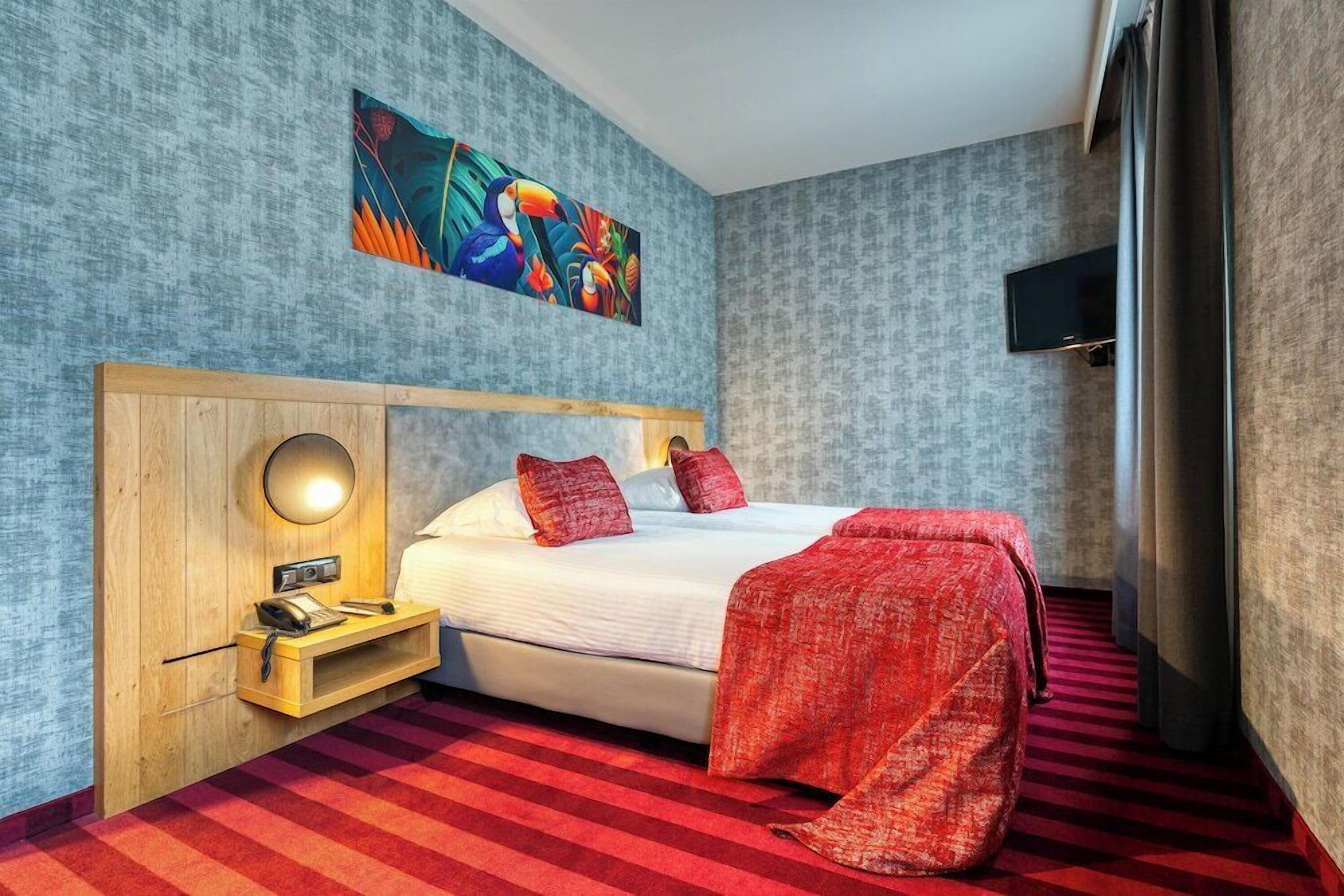 Habitación de hotel con cama, mesita de noche con lámpara, un cuadro en la pared y televisión también en la pared.