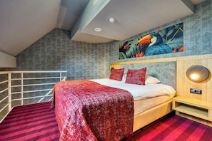 A bedroom with a bed, a red and black striped carpet, a wooden nightstand, a wall-mounted lamp, and a tropical-themed painting above the bed. - Hotel Restaurant Verviers (Verviers)