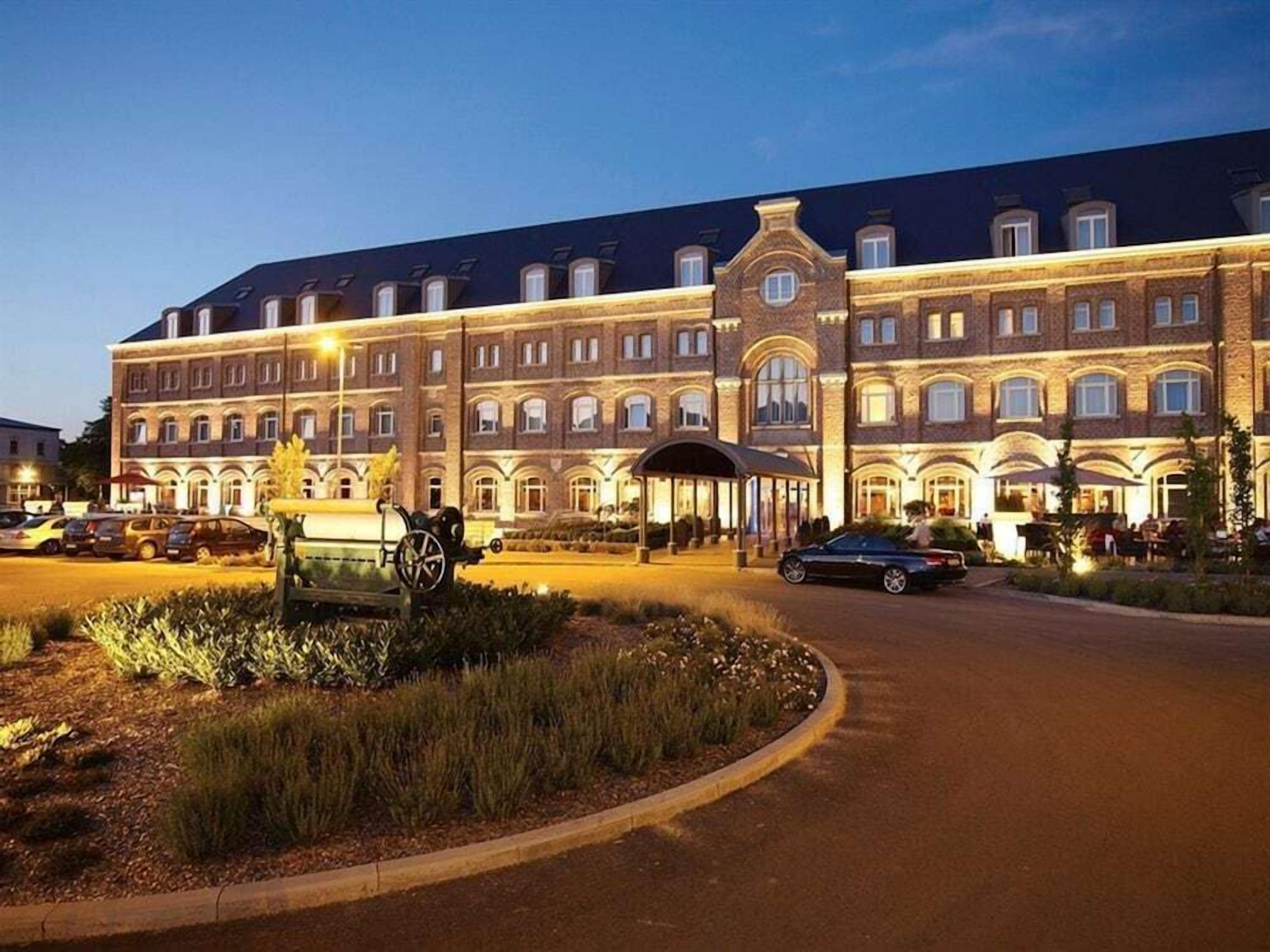 Un grand hôtel à plusieurs étages, de style architectural classique, avec une entrée principale dotée d’un passage couvert et un jardin bien entretenu.