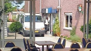 An outdoor seating area with tables and chairs, a brick building in the background, and a van parked outside.