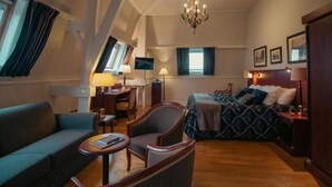 A hotel room with a bed, a sofa, a chair, a desk, a TV, and a chandelier. - Grand Hotel De Draak And Residence Dagmara (Bergen op Zoom)