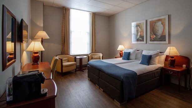 A hotel room with a bed, two bedside tables, a chair, a desk, a lamp, a mirror, and artwork on the wall. - Grand Hotel De Draak And Residence Dagmara (Bergen op Zoom)