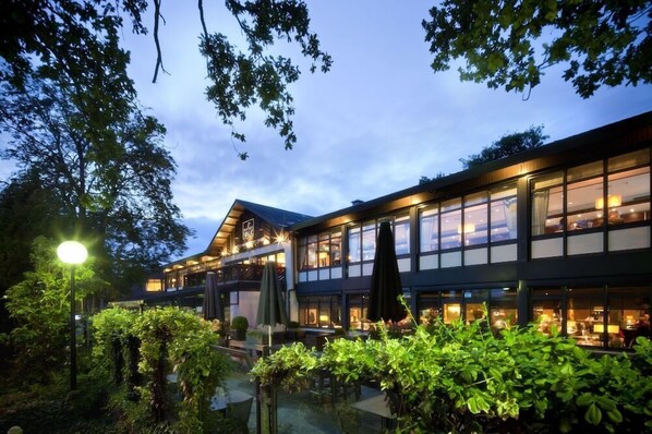 A modern hotel with a glass facade, outdoor seating, and lush greenery. - De Bilt - Utrecht (De Bilt)