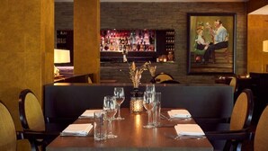 A restaurant with a bar in the background, a table set with glasses and a vase of flowers, and a painting on the wall.