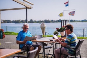 Dos personas sentadas en una mesa al aire libre con vista a un lago y banderas al fondo.