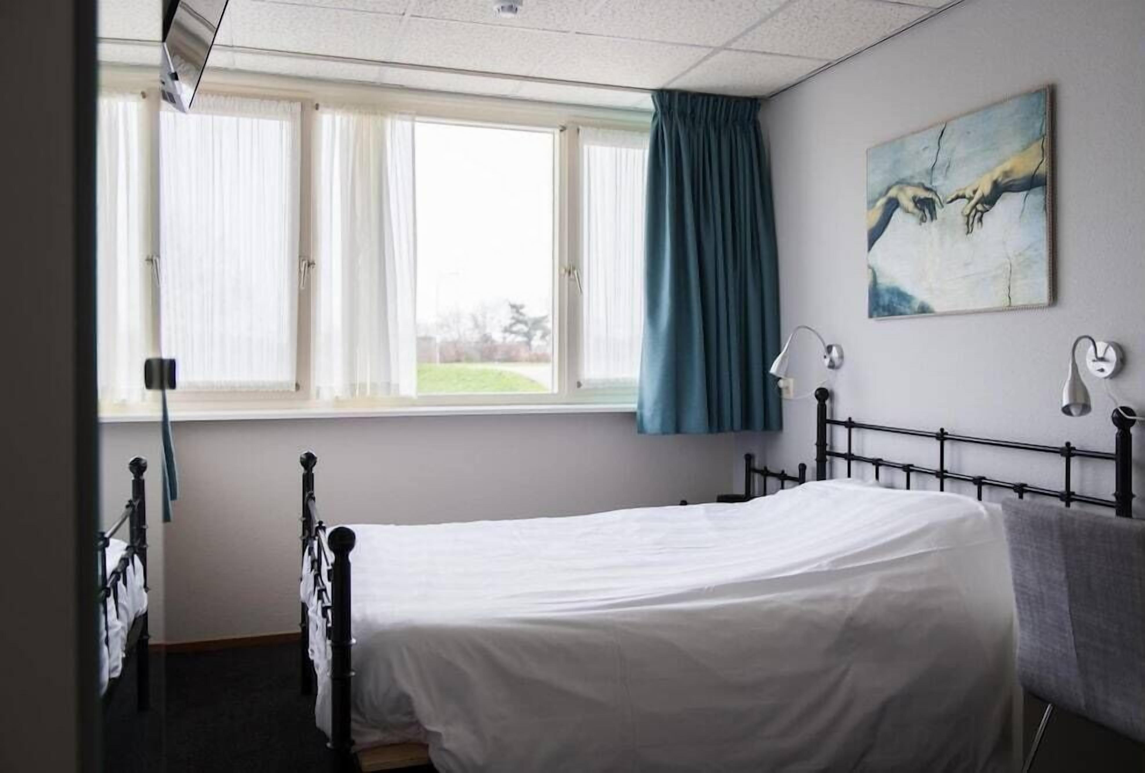A hospital room with a single bed, a window with curtains, a painting on the wall, and a bedside lamp.