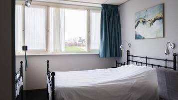 A hospital room with a single bed, a window with curtains, a painting on the wall, and a bedside lamp.