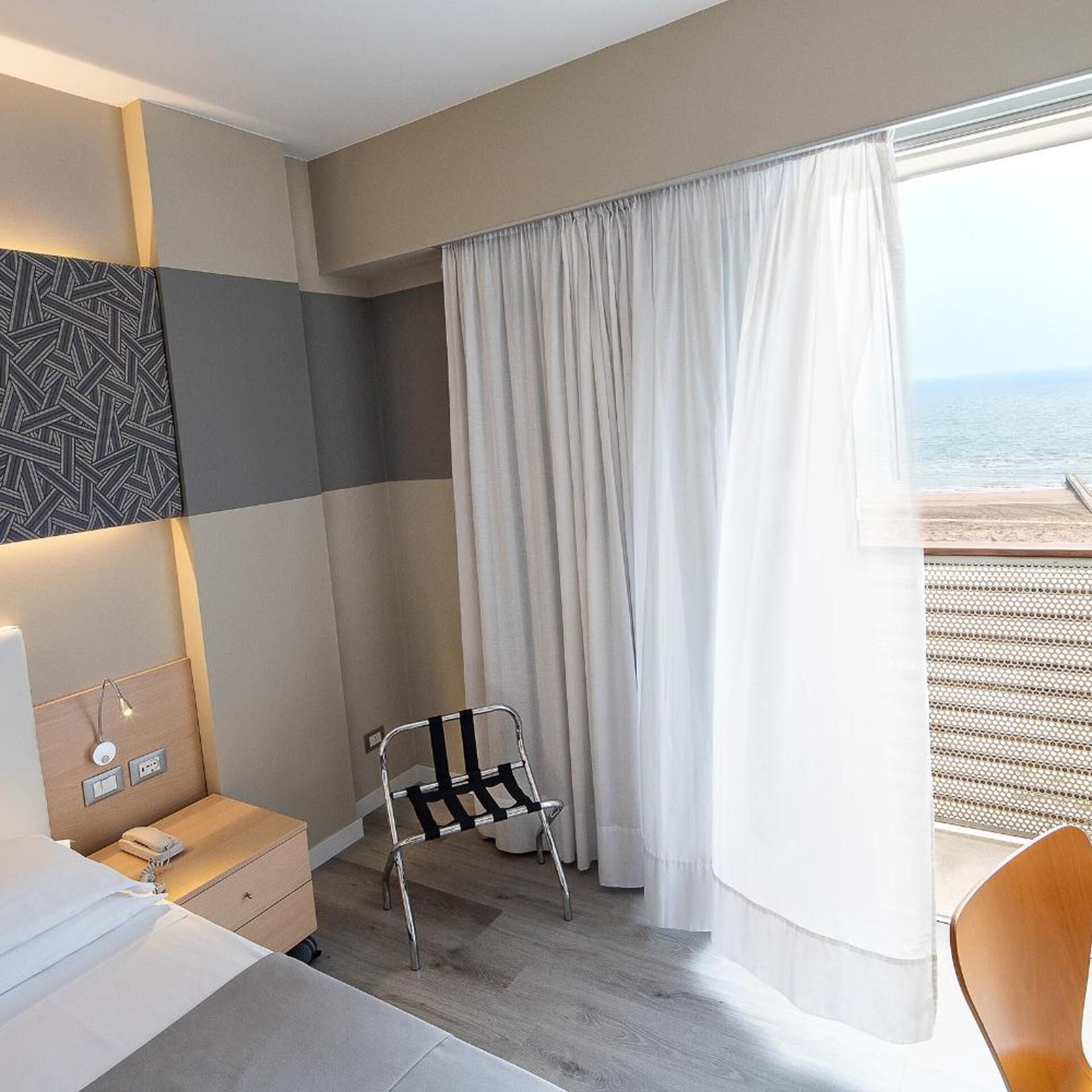 A hotel room with a bed, a chair, a desk, a window with curtains, and a view of the beach.