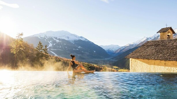 A person sitting in a clear pool with a mountain backdrop and a small building with a cross on top. - Hotel Bagni Vecchi (Valdidentro)