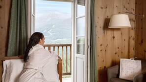 A room with a wooden paneled wall, a window with a view of a mountainous landscape, a chair, a lamp, and a bag on a table. - Hotel Bagni Vecchi (Valdidentro)