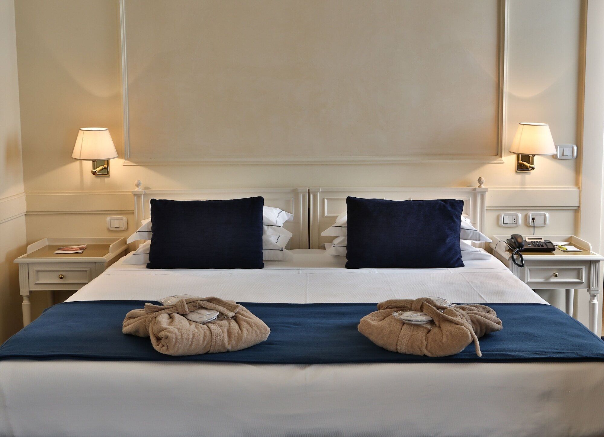 A hotel room with a large bed, two bedside lamps, a nightstand with a phone, and a wall-mounted light switch.